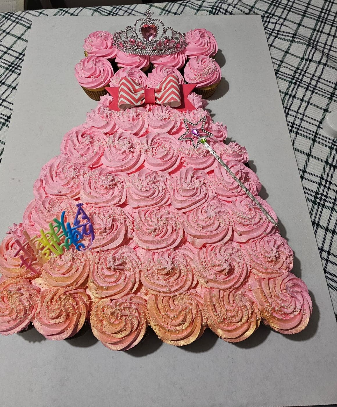 Pull-apart cupcake cake shaped like a pink princess dress, with swirled pink and peach frosting, pearl sprinkles, a chevron bow, a tiara, a wand and a Happy Birthday topper.