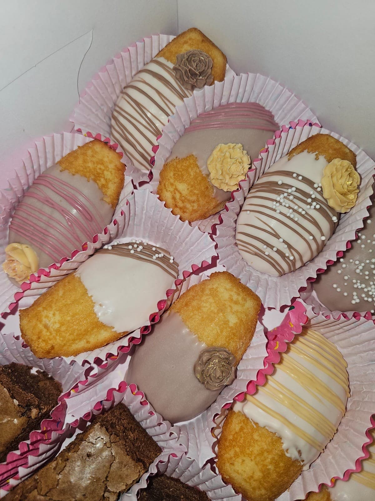 Golden sweet breads half-dipped in white, pink and chocolate coatings with drizzles, pearl sprinkles and sugar roses, beside brownies in pink cups.
