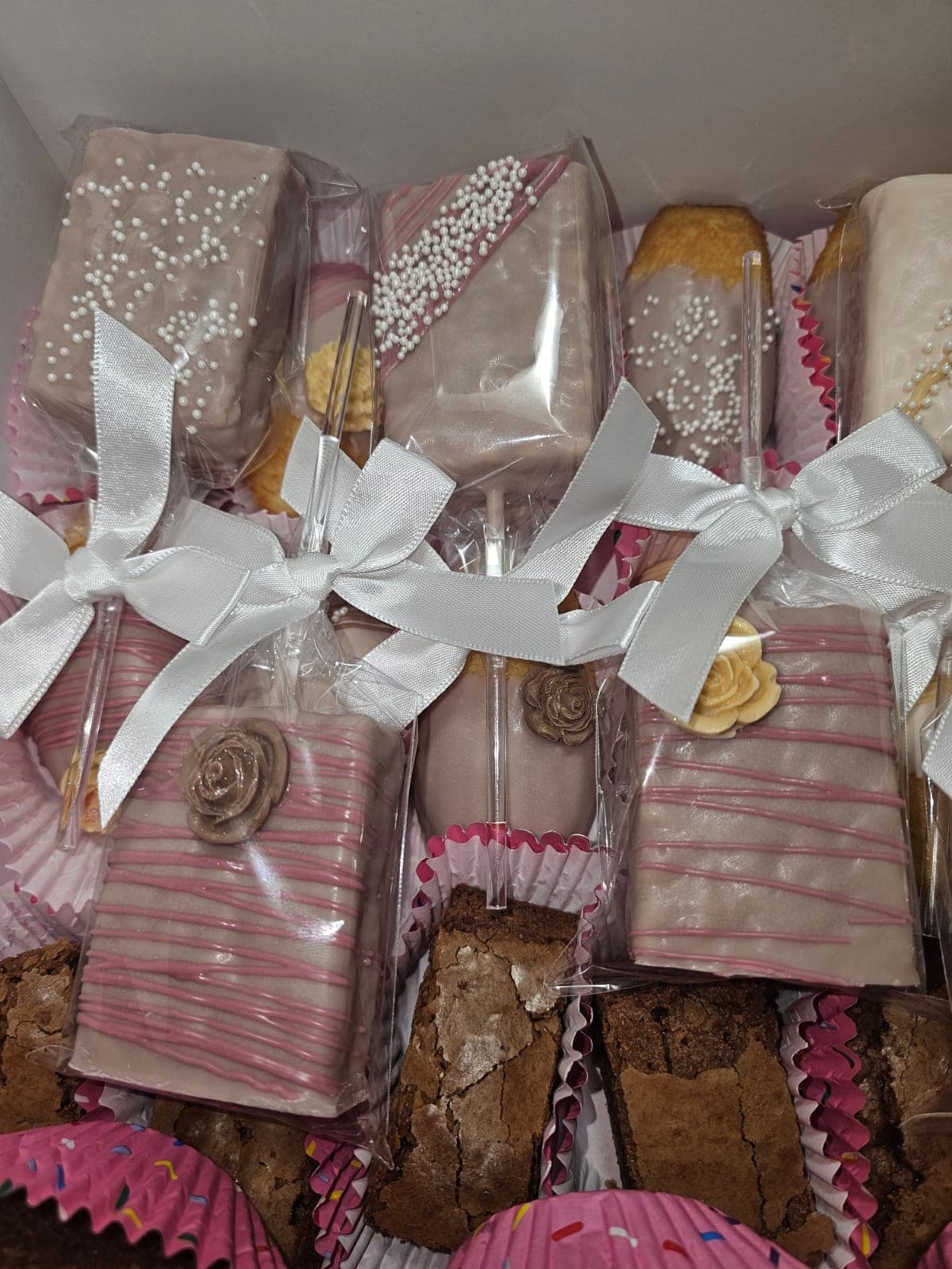 Chocolate-dipped treat pops with pink drizzle, pearl sprinkles and sugar roses, wrapped with white satin bows, next to brownies.