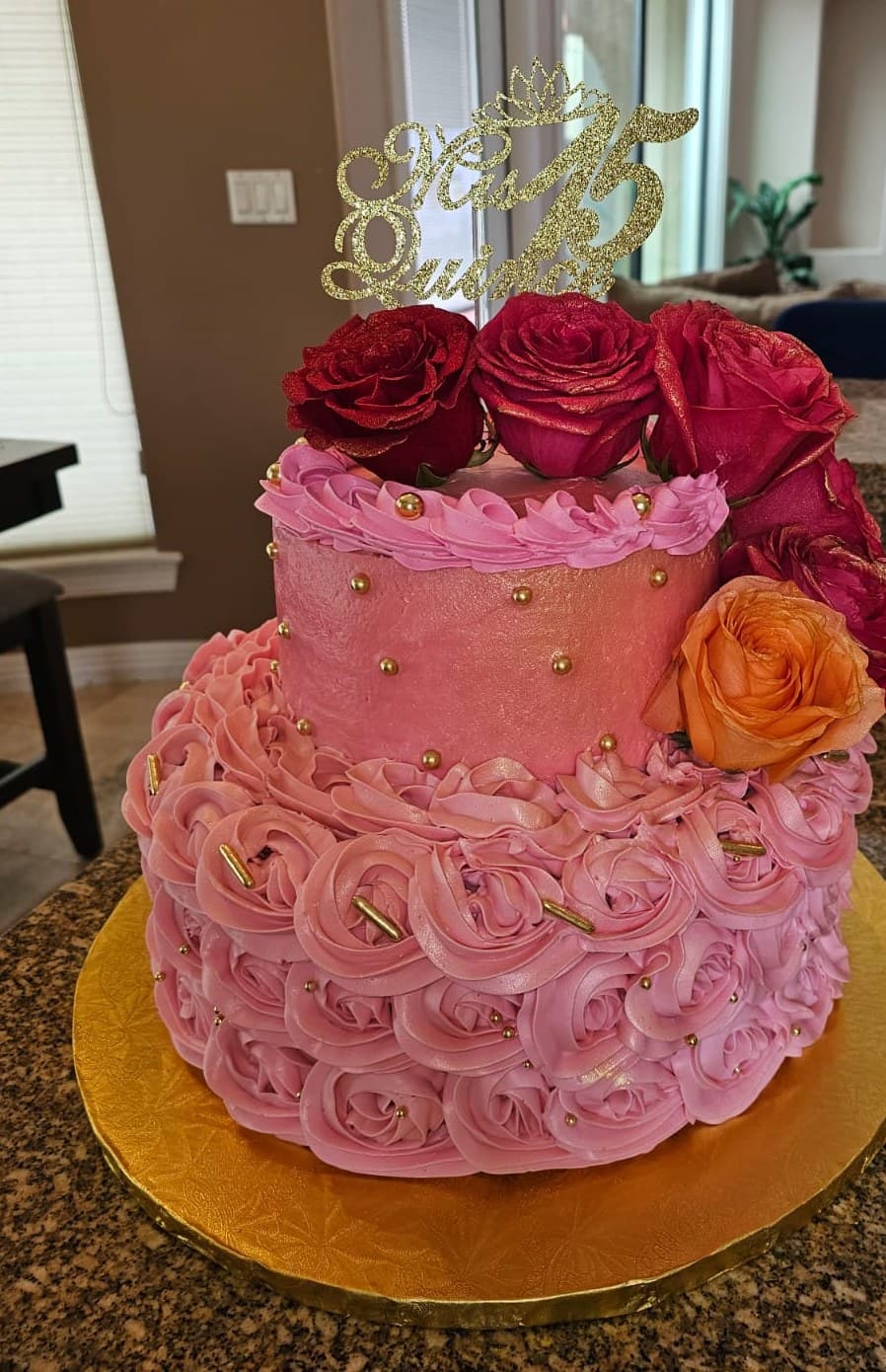 Pink two-tier quinceañera cake with buttercream roses, gold pearls, fresh red and orange roses and a gold “Mis 15” topper.