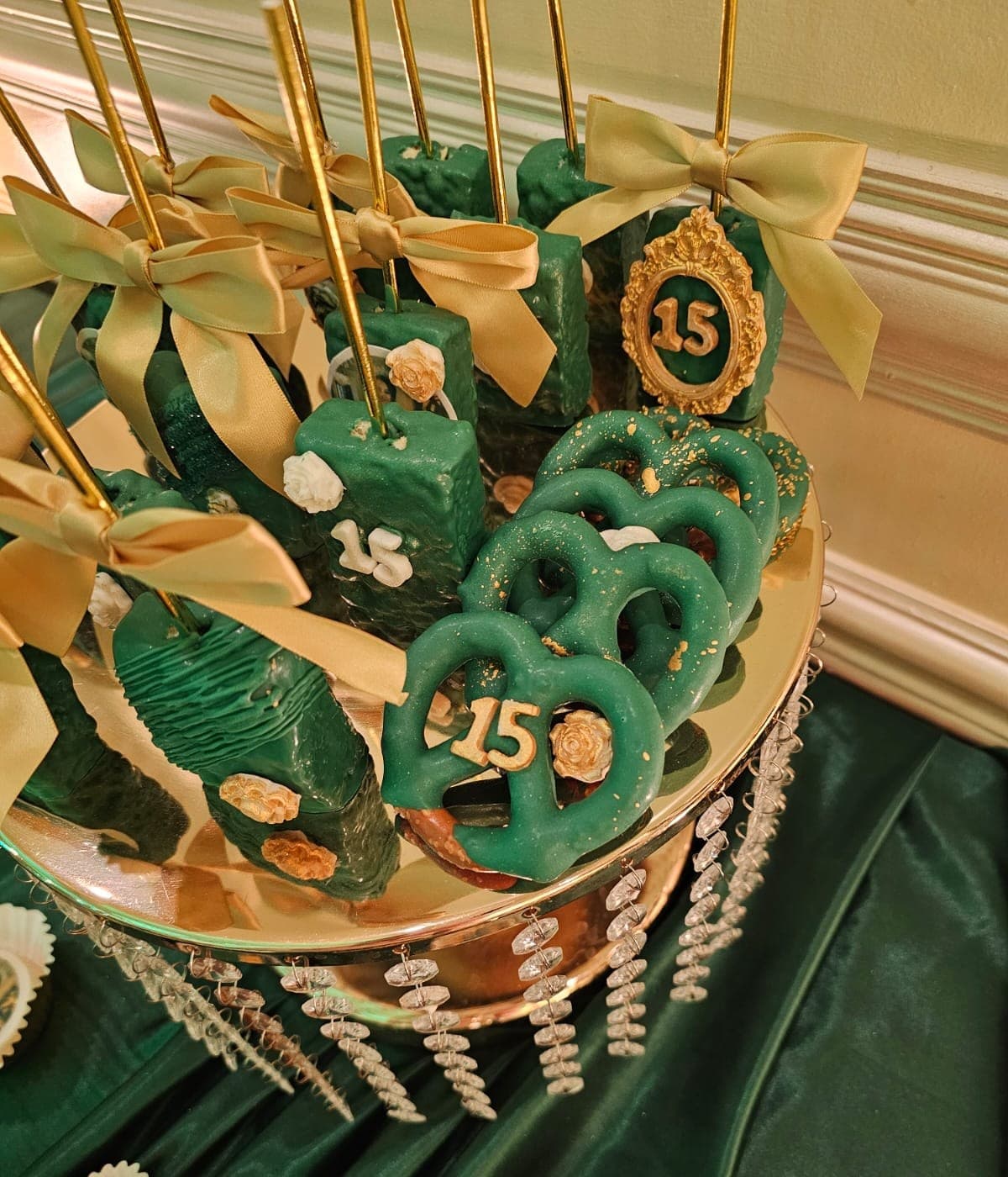 Emerald and gold quinceañera treats: dipped treat pops with gold bows and green pretzels with gold “15” toppers.